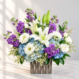 Purple and white floral arrangement with lilies and blue hydrangeas in a metallic vase
