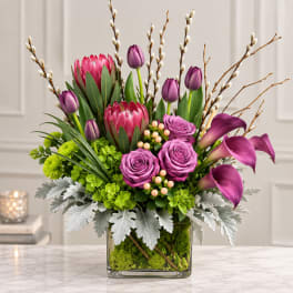 Mixed bouquet with pink protea, purple roses, calla lilies, and tulips in a glass vase