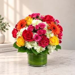Bright mixed bouquet of roses, gerberas, and hydrangeas in a green glass vase