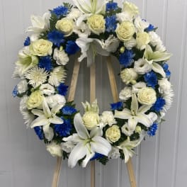 Circular funeral wreath with white lilies, cream roses, and blue flowers on a stand