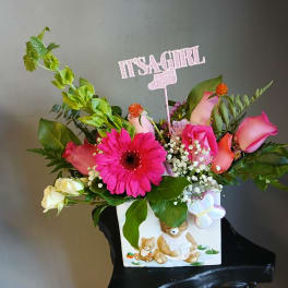 Pink and peach floral arrangement in a white teddy bear vase with a "It's a Girl" topper.