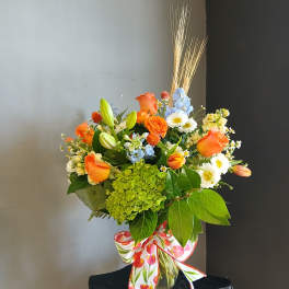 Mixed bouquet of orange roses, lilies, and daisies in a glass vase with a patterned ribbon