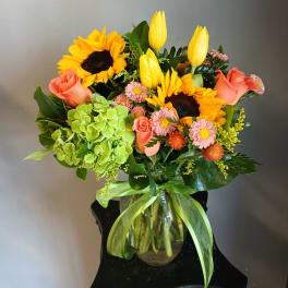Mixed bouquet in a glass vase with sunflowers, yellow tulips, pink roses, and green hydrangea.