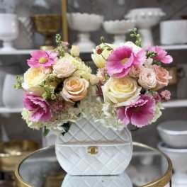 Handbag-shaped white ceramic vase filled with pink anemones, pastel roses, and white hydrangeas on a gold table