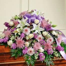 Lavender and white casket spray with lilies, roses, mums, and a purple bow on a wooden casket.