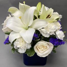 Compact arrangement of white lilies, roses, and carnations with purple accents in a dark blue cube vase
