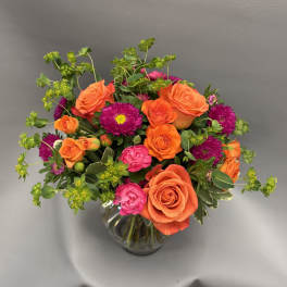 Bright arrangement of orange roses, pink blooms, and purple asters in a clear glass vase