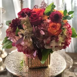 Compact bouquet of red, orange, and pink flowers in a gold vase