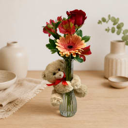 Bouquet of red and peach flowers in a glass vase with a teddy bear