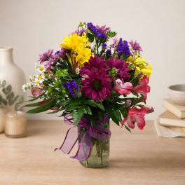 Mixed bouquet of daisies, lilies, and purple flowers in a glass vase with ribbon