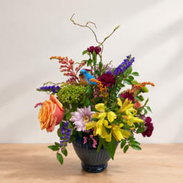 Colorful mixed flower arrangement in a dark vase with a small blue bird ornament
