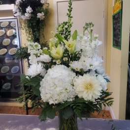 Tall white floral arrangement with lilies, hydrangeas, and daisies in a glass vase
