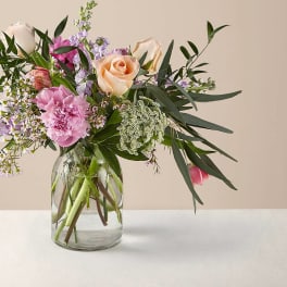 Loose arrangement of pink peonies, peach roses, and pastel blooms in a clear glass jar vase