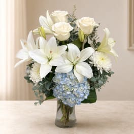 White lilies, roses, and blue hydrangeas arranged in a clear glass vase