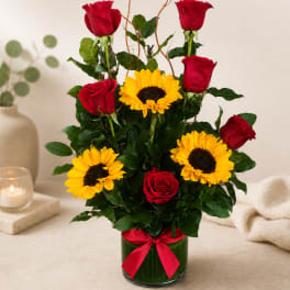 Red roses and yellow sunflowers arranged in a glass vase with a red ribbon