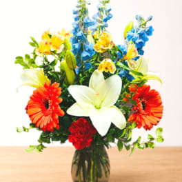 Mixed bouquet with white lilies, orange gerbera daisies, blue delphinium, and red carnations in a glass vase