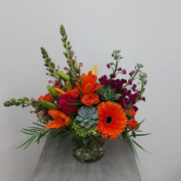 Bright mixed arrangement with orange gerbera, lilies, roses, and succulents in a clear glass vase