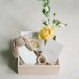 Gift box with a yellow flower, baby book, plush bunny ears, and a card