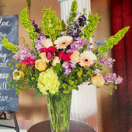 Tall mixed arrangement of pink, peach, and green flowers in a clear glass vase on a table