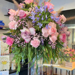 Tall bouquet of pink roses, lavender flowers, and blue blooms in a clear glass vase.