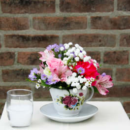 Pink and purple flowers arranged in a teacup on a saucer beside a white candle.