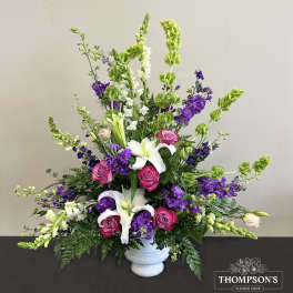 Tall fan-shaped arrangement with white lilies, pink roses, purple blooms, and green bells of Ireland in a white urn.