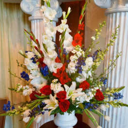 Tall red, white, and blue floral arrangement with roses, lilies, and delphinium in a white urn vase