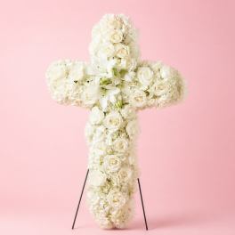 White floral cross arrangement on a stand against a pink background