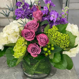 Round bouquet of mauve roses, purple stock, and white hydrangeas in a clear glass vase