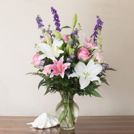 Tall arrangement of white lilies, pink roses, and purple flowers in a clear glass vase.