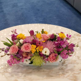 Mixed pink, purple, and yellow floral arrangement in a low white container