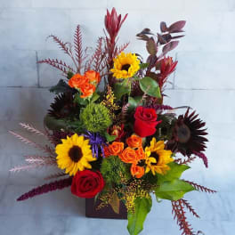 Mixed bouquet with sunflowers, roses, and orange spray roses in a square vase