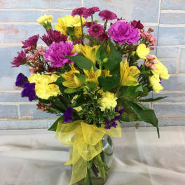 Mixed bouquet of yellow, pink, and purple flowers in a glass vase with a yellow ribbon