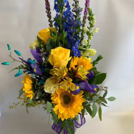 Tall mixed bouquet with sunflowers, yellow roses, and blue flowers in a glass vase
