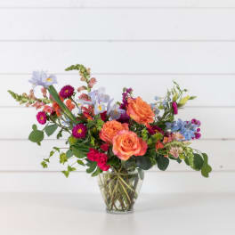 Mixed bouquet of orange, pink, and blue flowers in a clear glass vase
