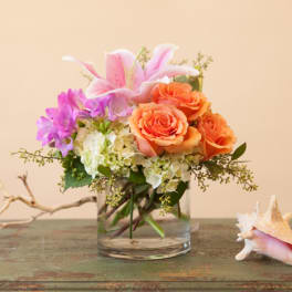 Pink lilies and orange roses in a clear glass vase with a seashell beside it.