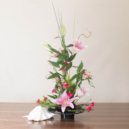 Ikebana-style arrangement of pink lilies and carnations in a low black dish with a white seashell accent.
