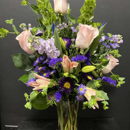 Tall vase arrangement of pink roses and lilies with purple flowers and green spikes.