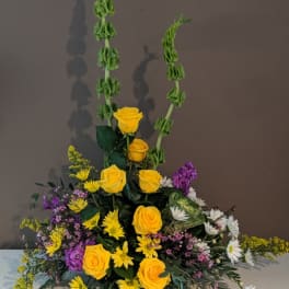 Tall floral arrangement with yellow roses, daisies, and purple accents