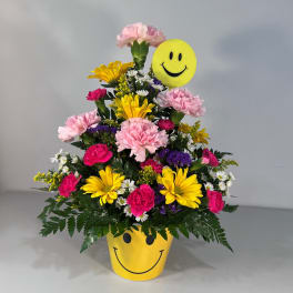 Smiley-face flower arrangement in a yellow pot with a matching smiley topper
