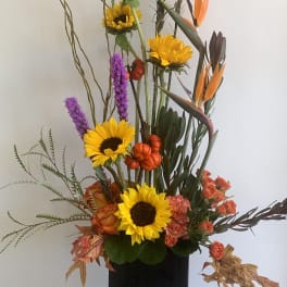 Tall floral arrangement with sunflowers, orange blooms, and purple spikes in a black vase