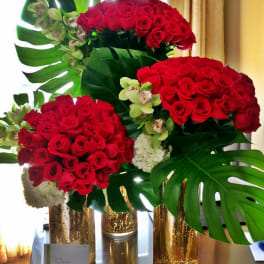 Red rose arrangements with orchids and large tropical leaves in gold vases