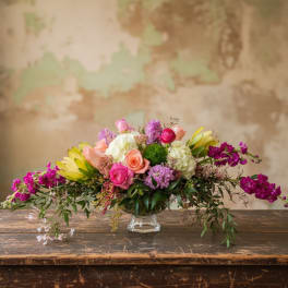 Low centerpiece of pink and peach roses with white hydrangeas, ranunculus and magenta stock in a clear glass bowl.