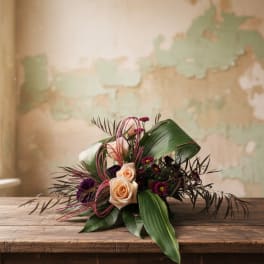 Low arrangement of peach roses and dark purple blooms framed by large green leaves on a rustic wooden table