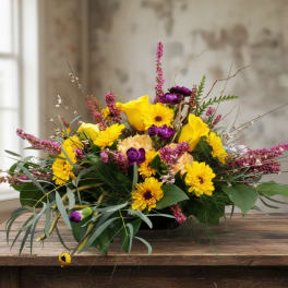 Low arrangement of yellow gerbera and calla lilies with purple blooms and pink spikes in a shallow container