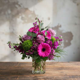 Bright pink gerbera daisies, lavender roses, and green dianthus in a clear square glass vase