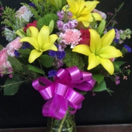 Bouquet of yellow lilies, pink flowers, and a purple ribbon in a glass vase