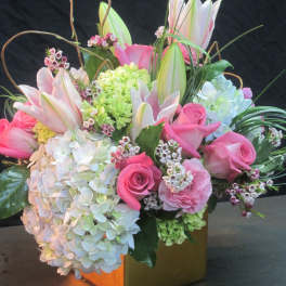 Compact arrangement of pink roses, white hydrangeas, and lilies in a gold cube vase