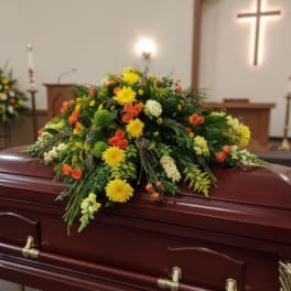 Casket spray of yellow, orange, and white flowers on a polished mahogany casket in a chapel.