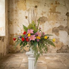 Tall mixed arrangement of pink lilies, red and yellow roses, and purple asters with ferns in a white urn on a pedestal.